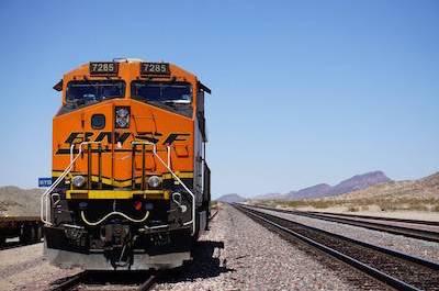 An orange train speeding through desert tracks, surrounded by vast sandy dunes under a clear blue sky.