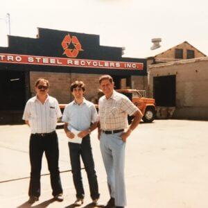 Three men in front of a metal recycling facility, wearing safety vests and hard hats, discussing operations.